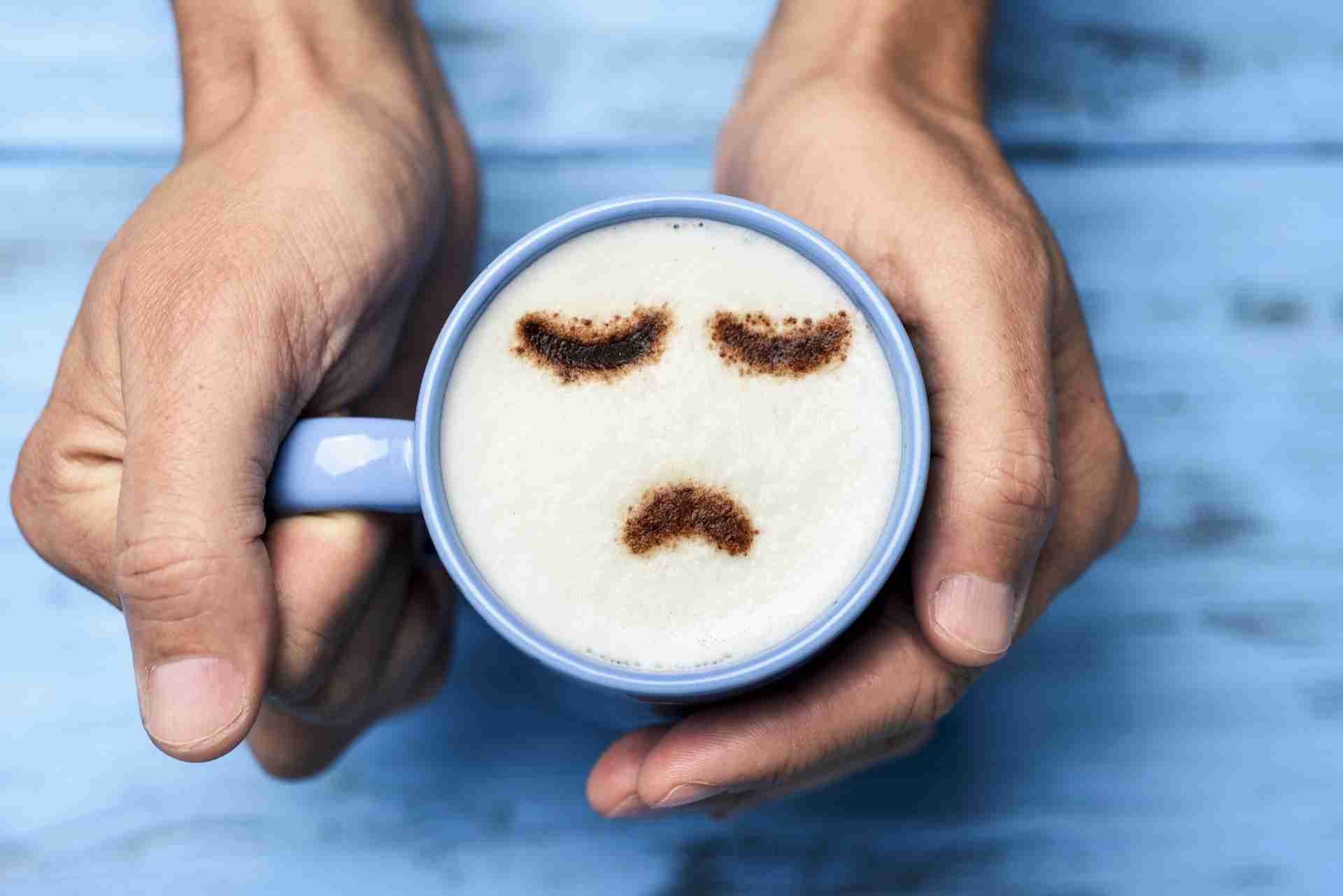 Hands holding a latte with a sad face drawn in the foam, symbolizing depression