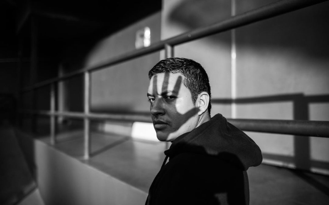 Black and white portrait of a man in a hoodie glancing back, a shadow falling across his face suggesting stigma and isolation