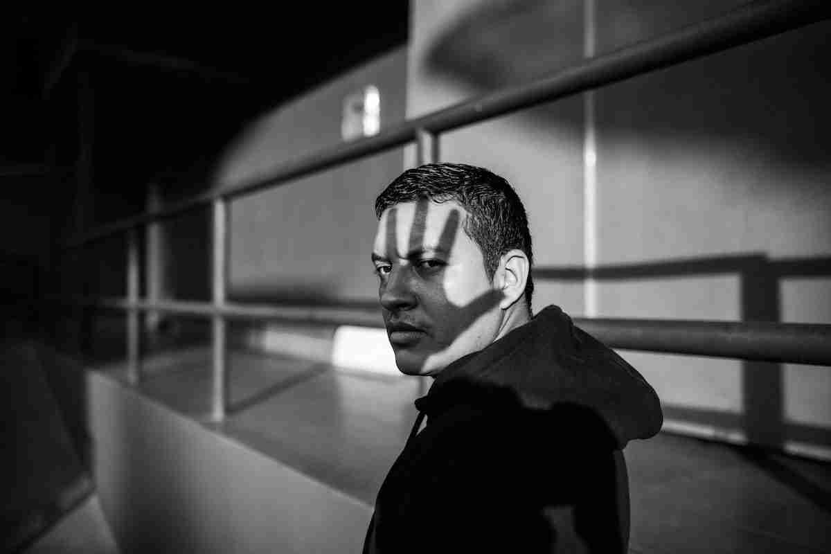 Black and white portrait of a man in a hoodie glancing back, a shadow falling across his face suggesting stigma and isolation